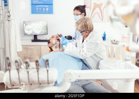 Stomatologue senior femme avec masque de protection effectuant une inspection dentaire après avoir mis l'implant dans la bouche de l'homme malade dans la clinique orthodontique. Médecine dentaire médecin traitant les dents du patient Banque D'Images