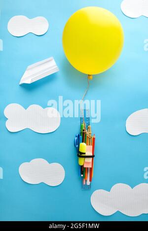 Ballon jaune levant un bouquet de fournitures scolaires avec fond bleu et découpe de nuages. Concept de retour à l'école. Composition verticale. Banque D'Images