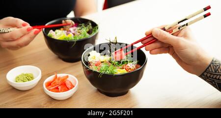 Homme et femme mangeant de la salade de poke avec des baguettes. Verser la salade de thon dans un bol. Les gens du restaurant mangent de la salade avec des baguettes. Salade de fruits de mer asiatiques Banque D'Images