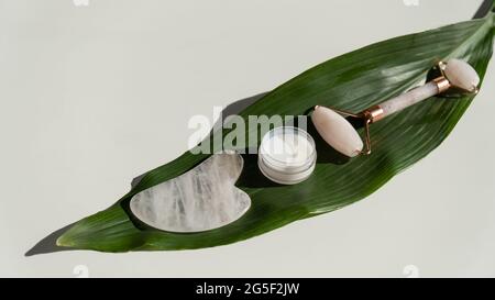 Grattoir de gouache rose, rouleau, pot de crème de feuille verte sur une table blanche. Équipement pour l'auto-massage et les soins de la peau pour le visage et le cou Banque D'Images