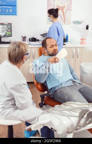 Stomatologue senior femme finissant la chirurgie dentaire pour homme patient assis sur une chaise de stomatologie regardant la dent. Médecin expliquant le traitement des maux de dents pendant la consultation stomatologique Banque D'Images