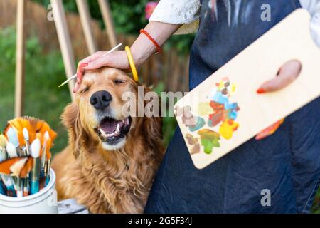 Jeune artiste femme travaillant sur son art toile peinture à l'extérieur dans son jardin avec Golden retriever gardant son entreprise. Concept de loisirs créatifs. Banque D'Images