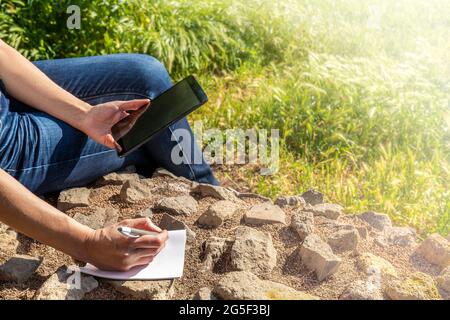 Gros plan des mains d'une fille prenant des notes tout en utilisant son smartphone pendant une promenade à la campagne. Banque D'Images