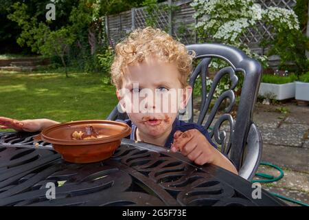 Un blanc collant petit garçon caucasien âge près de 3 ans avec des cheveux blond bouclés est assis à l'extérieur avec une bouche marron groubby manger gâteau au chocolat Banque D'Images