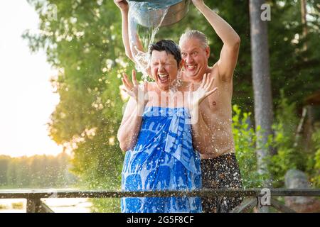 Un couple âgé s'est amusé après un sauna finlandais sur une jetée en bois dans un lac. Homme mature versant de l'eau froide sur sa femme. Été typiquement finlandais. Banque D'Images