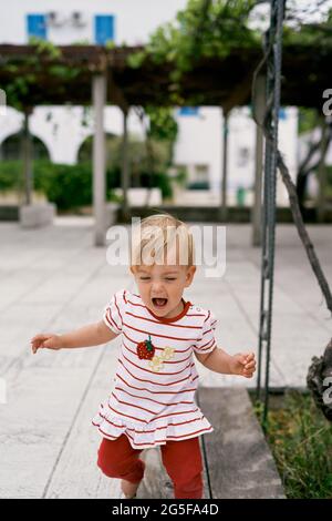 Une petite fille marche pieds nus dans la cour de la maison sur fond de pergola et de verdure Banque D'Images