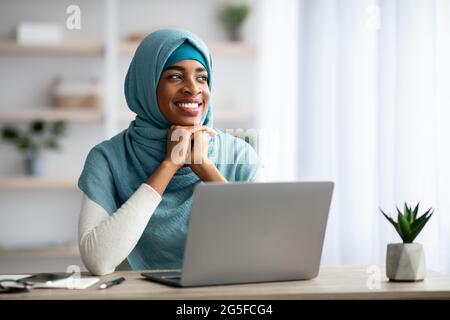 Portrait de la jeune fille musulmane noire souriante dans le hijab assis au bureau avec ordinateur et regardant loin, la dame musulmane africaine rêveuse dans le foulard Enjoyin Banque D'Images