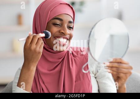 Belle femme musulmane noire dans hijab regardant dans miroir grossissant et appliquant le blush à la maison, heureuse femme islamique africaine tenant le maquillage Brush et SM Banque D'Images