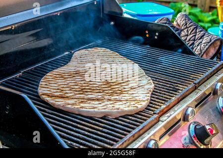 Cuisine de pain plat maison rustique sur un grill extérieur avec des gants de four et de la vaisselle en plastique floue sur la table à côté - attention sélective Banque D'Images