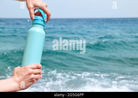 Bouteille thermique en acier dans les mains de la femme dans une journée ensoleillée d'été sur le fond bleu de la mer. Mode de vie sain, contrôle de l'équilibre de l'eau, concept de soins de la peau Banque D'Images