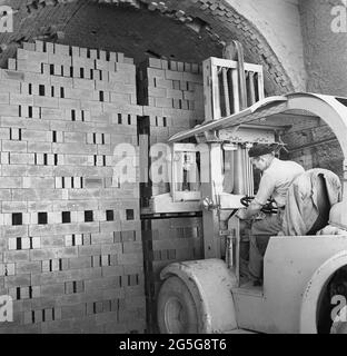 Années 1950, ouvrier historique et masculin exploitant un chariot élévateur électrique pour déplacer une grande pile de briques, Bedford, Angleterre, Royaume-Uni. Le déclenchement de la première Guerre mondiale a conduit au développement de machines avec des plates-formes montantes et abatrices et le premier chariot élévateur est considéré comme un véhicule appelé tracteur de camion fabriqué en 1917. Les développements des années 1940 ont vu des chariots élévateurs introduits avec une batterie rechargeable afin qu'ils puissent être utilisés toute la journée. Banque D'Images