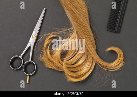 Une serrure de cheveux blonds courbés de femme, des ciseaux et un peigne sur un fond sombre, vue du dessus Banque D'Images