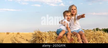 Deux adorables frères et sœurs caucasiens aiment s'amuser assis sur le dessus de la balle de foin doré sur le champ de blé récolté près de la ferme. Bonne enfance et Banque D'Images