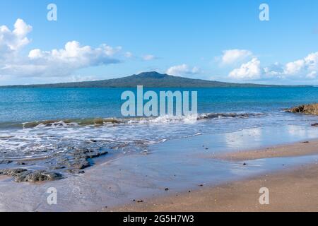 De petites vagues se brisent sur la rive avec l'île Rangitoto à l'horizon lointain en face du port de la plage de St Leonard, North Shore Auckland. Banque D'Images