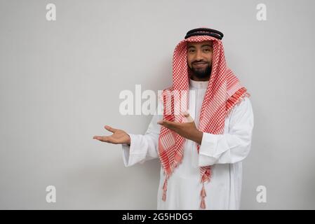 un jeune homme arabe dans un turban souriant tout en regardant la caméra avec un geste de main présentant quelque chose Banque D'Images
