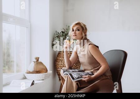 Jeune femme d'affaires travaille à la maison.pensive modèle calme tient un verre de vin blanc et un magazine et regarde la caméra. Belle femme dans un I moderne Banque D'Images