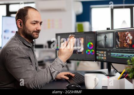 Éditeur vidéo parlant sur vidéocall avec le client tenant le smartphone éditant le film assis dans l'agence de création. Vidéaste traitant le montage de film en société multimédia avec logiciel de post-production Banque D'Images