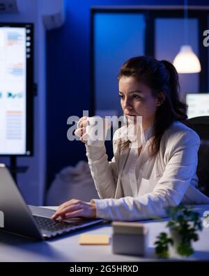 Femme indépendante avec une tasse de café tapotant sur l'informatique d'affaires surtravaillant au projet de marketing. Entrepreneur très occupé utilisant le réseau de technologie moderne sans fil tard dans la nuit dans le bureau de l'entreprise Banque D'Images