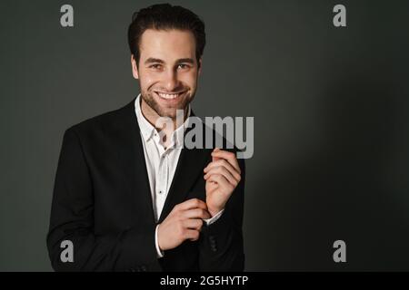 Un homme brunette non rasé en veste souriant et regardant la caméra isolée sur fond noir Banque D'Images