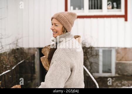 Femme souriante portant un chapeau tricoté portant du bois de chauffage tout en regardant loin Banque D'Images