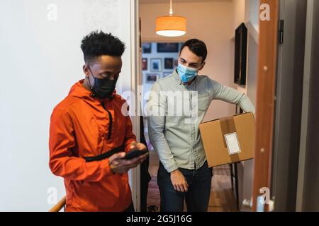 Homme de taille moyenne avec colis debout par un liveur de sexe masculin utilisant un téléphone intelligent pendant une pandémie Banque D'Images