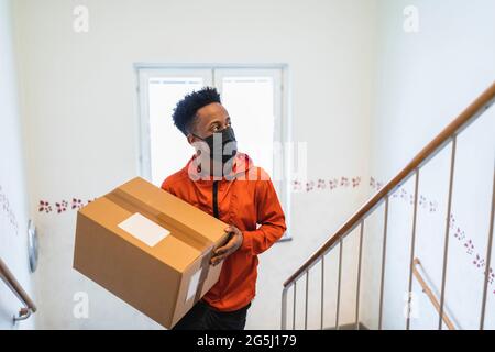 Jeune liveur transportant le colis tout en se déplaçant dans l'escalier pendant une pandémie Banque D'Images