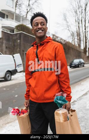 Jeune liveur heureux transportant des sacs en papier tout en marchant sur la route Banque D'Images