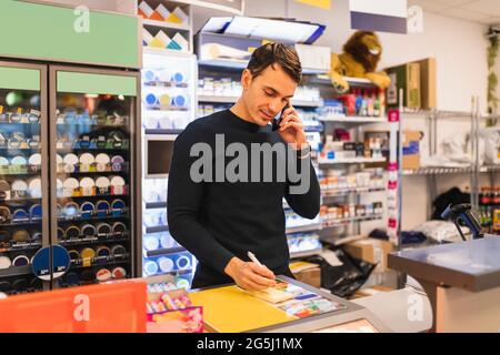 Un homme écrit par le propriétaire d'un magasin en parlant sur un téléphone portable au moment de la caisse Banque D'Images