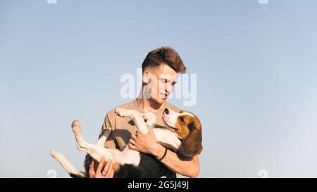 Le jeune homme tient son chien de beagle heureux dans les mains, tiré contre le ciel bleu clair. Style de vie avec les animaux de compagnie, heureux propriétaire de chiot, chien de race Banque D'Images