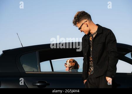 Le jeune homme se tient à côté de la voiture, le chien de beagle regardant par la fenêtre de la voiture. Mode de vie avec animaux, concept voyager avec chien Banque D'Images
