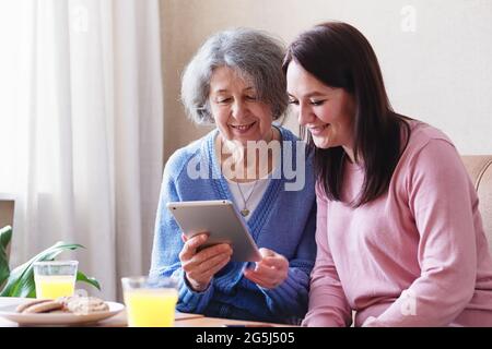 La fille rend visite à une mère âgée et utilise une tablette ensemble pour voir les réseaux sociaux - une femme âgée et une jeune femme sont assis à la maison sur le canapé A. Banque D'Images