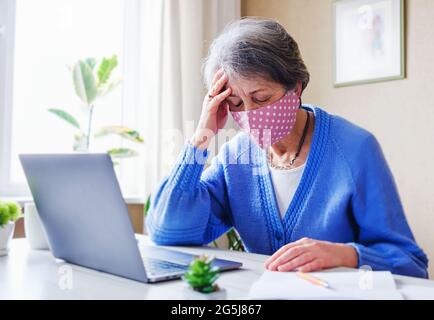 Une femme âgée dans un masque avec un ordinateur portable subit le stress et les maux de tête dans le cadre de la pandémie du coronavirus - UN pensionné portant un masque facial utilise un co Banque D'Images