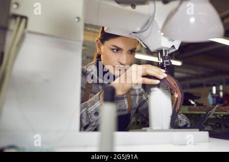 Jeune femme concentrée regardant l'aiguille tout en travaillant sur la machine à coudre à l'usine de chaussures Banque D'Images