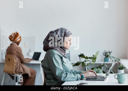 Portrait en vue latérale d'une femme moderne du Moyen-Orient portant un foulard au bureau et utilisant un ordinateur portable, espace de copie Banque D'Images