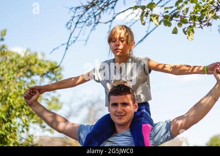 Frère défoqué de la sœur de circonscription à l'arrière. Portrait d'une fille heureuse sur les épaules de l'homme, porcgyback. Une fille vole, lève les mains. Famille jouant à l'extérieur. Bleu vert Banque D'Images