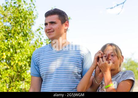 Défocused souriant préteen caucasienne fille jouant avec le doigt jeune homme caucasien. Été extérieur ciel bleu et vert arbre fond. Imbécile. Sortie Banque D'Images