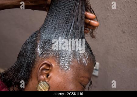 Coiffeur relaxant les cheveux sur la tête d'une femme africaine et également en utilisant le peigne pour étirer et appliquer la crème relaxante à travers les cheveux Banque D'Images