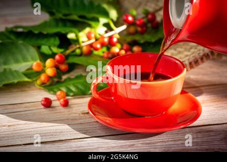 Le café est versé dans une tasse rouge avec des graines de café et des feuilles sur une table en bois. Banque D'Images