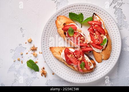 Sandwichs ouverts avec fraises, fromage doux à la menthe et aux noix dans une assiette en céramique sur fond de pierre gris clair. Nourriture estivale et saine, légumes Banque D'Images