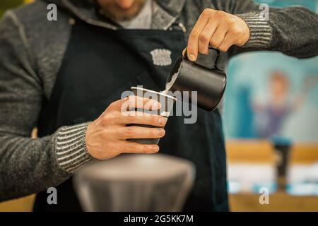 Barista méconnaissable portant un tablier noir pour préparer un cappuccino, versant du lait dans une tasse en papier dans le bar à café Banque D'Images