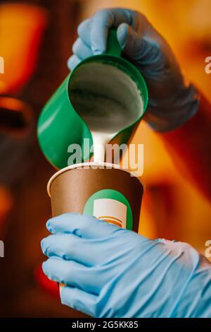 Gros plan des mains tenant le barista professionnel qui verse du lait chaud dans une tasse de papier à café. Préparation du café, concept de service Banque D'Images