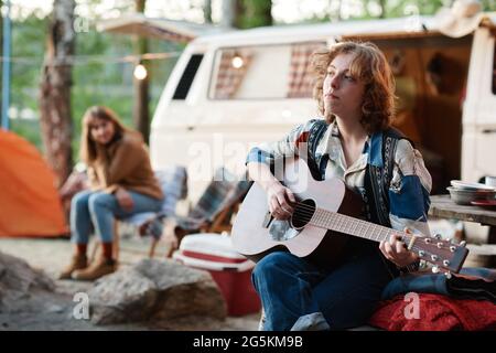 Jeune femme assise à l'extérieur et jouant de la guitare pendant le camping dans la forêt Banque D'Images