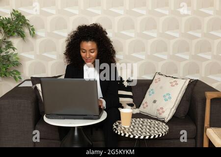 Femme noire concentrée assise dans un canapé dans le bureau et utilisant son ordinateur portable. concept de bureau. Banque D'Images