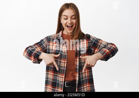 Image de la femme caucasienne souriante regardant, pointant vers le bas avec l'expression du visage heureux étonné, montrant cool nouvelle publicité, produit de logo, blanc Banque D'Images