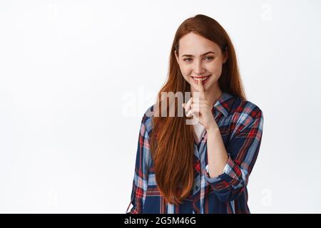 Image de jolie fille caucasienne avec des sourires de cheveux rouges, faire shush, appuyer le doigt sur les lèvres, calme ou tabou geste, se précipitant avec le visage mystérieux, debout Banque D'Images