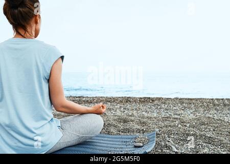 Une femme assise dans lotus pose pratique le yoga sur la plage le matin, elle médite, regardant sur la mer bleue. Concept de bien-être. Banque D'Images