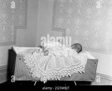 1960, historique, dans un coin d'une pièce, un bébé reposant sur un petit jeté décoratif tricoté sur un lit de voyage léger de l'époque, Angleterre, Royaume-Uni. Banque D'Images