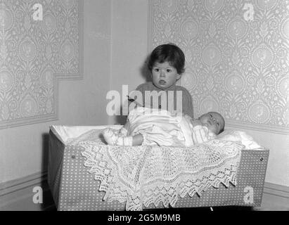 1960s, historique, dans un coin d'une pièce, une petite fille debout à côté d'un bébé reposant sur un jet de dentelle au-dessus d'un berceau de voyage de l'époque, Angleterre, Royaume-Uni. Banque D'Images