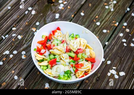 Salade de pâtes tortellini sur une terrasse en bois de table avec pétales de cerisier tombés sur le sol et tranches de poivron de concombre dans un bol Banque D'Images