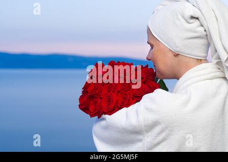 Une heureuse femme en robe blanche avec un bouquet de roses rouges donne sur le balcon de l'hôtel, au bord de la mer Banque D'Images
